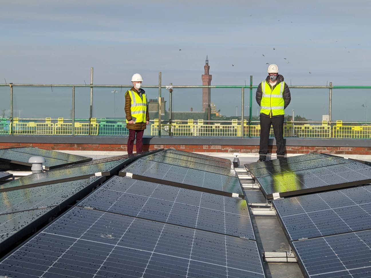 two people standing on a roof next to solar panels with grimsby's skyline in the distance