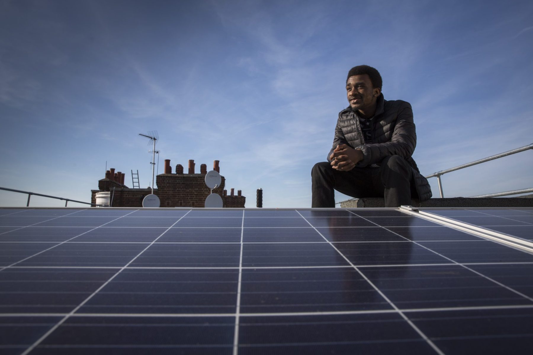 Photo of Ziggy, one of the Re-Powering interns and an estate resident, sat with the Solar photo voltaic (PV) panels on the roof of Hackney council estate Bannister House.