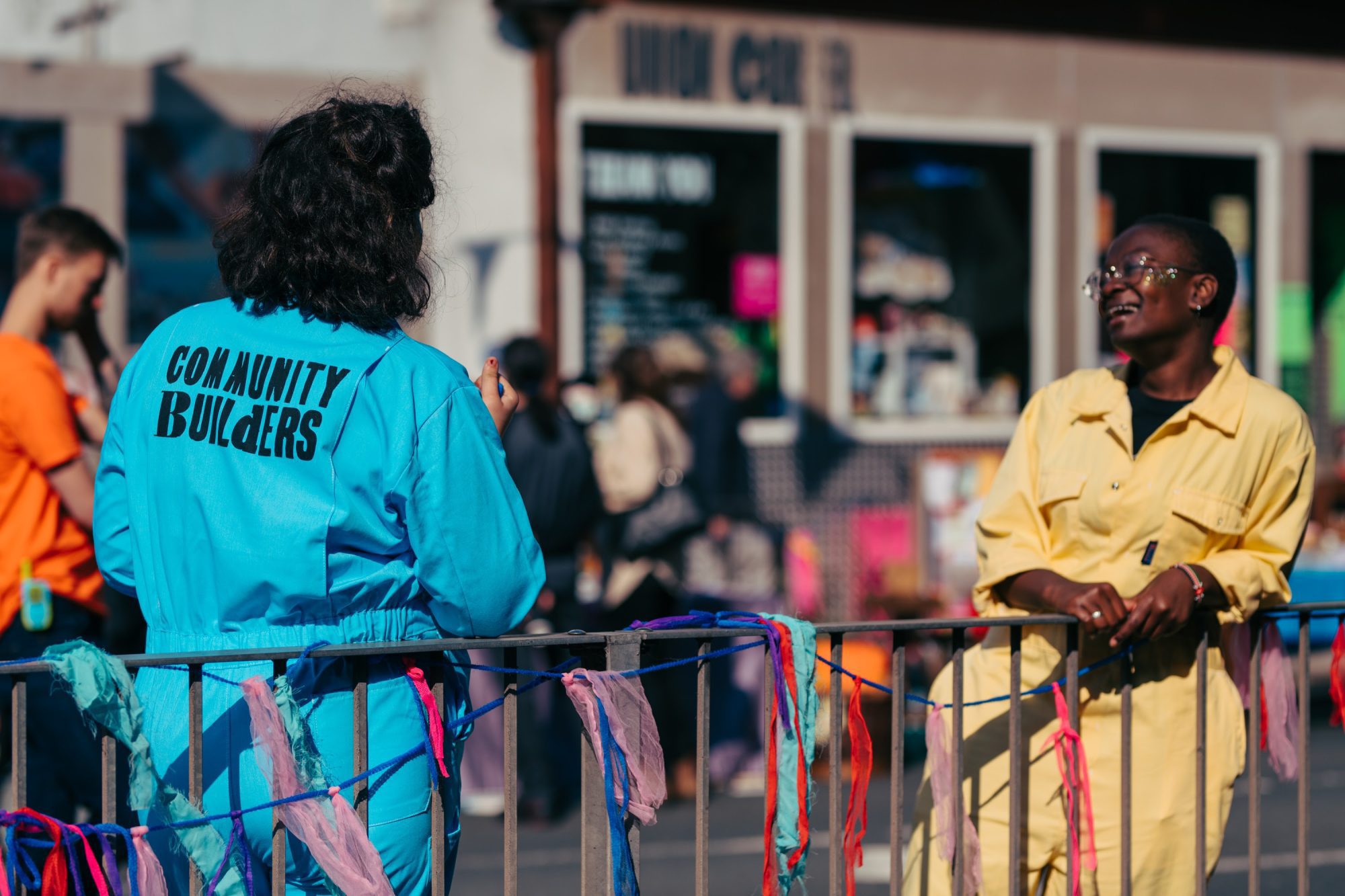 Two people in bright color jumpsuits that say 'Community Builders' talk at the Nudge Union Street block party.